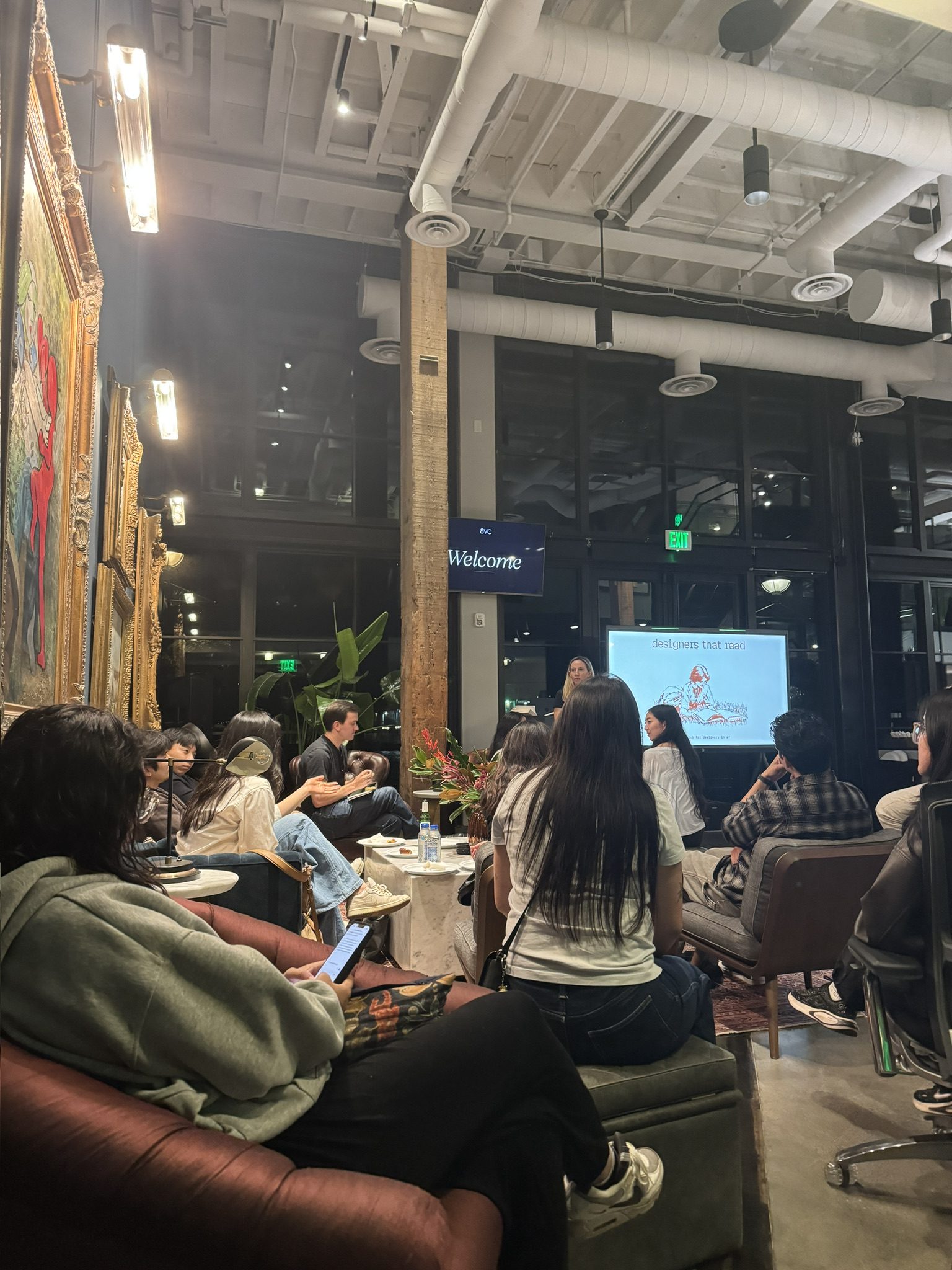 Attendees seated on couches and ottomans listening to a talk, with a screen in the background reading &ldquo;designers that read.&rdquo;