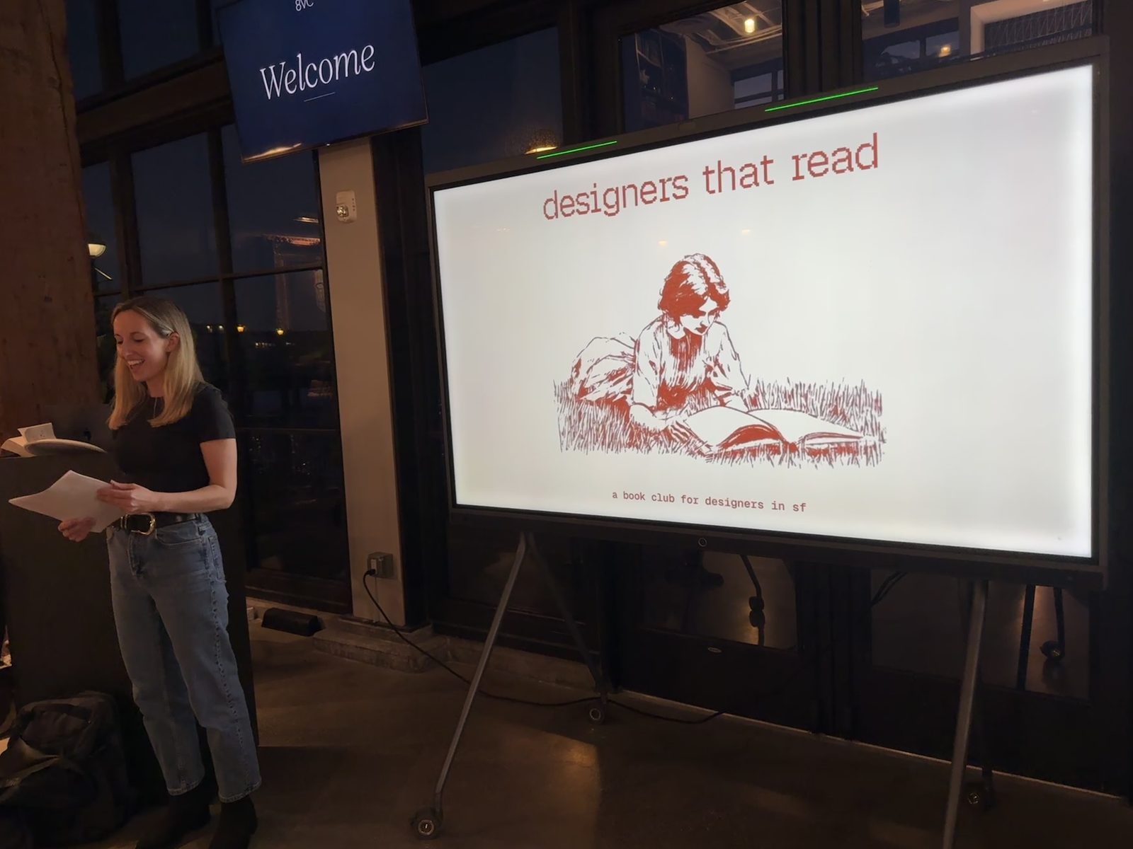 A woman speaking at a podium beside a large screen displaying the Designers that read logo and the tagline &ldquo;a book club for designers in sf.&rdquo;